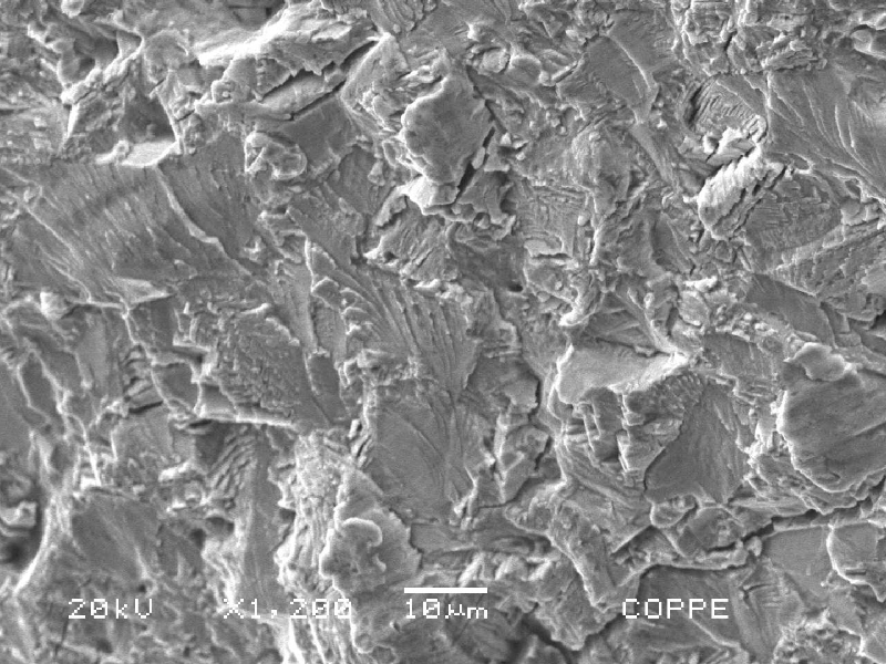 SEM fractograph at 1200x magnification showing an intergranular fracture surface with angular, faceted grains and fine feather-like markings.
