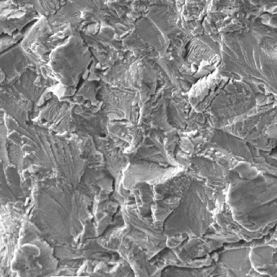 SEM fractograph at 1200x magnification showing an intergranular fracture surface with angular, faceted grains and fine feather-like markings.