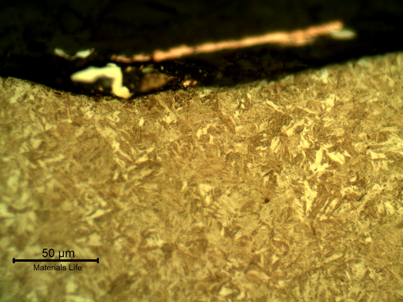 Micrographic detail of the plate section, close to the surface that suffered the impact.  After etching the lead alloy film at the surface appears dark gray and the copper alloy film appears clear and orange. No damage in the plate martensitic microstructure at this magnification.