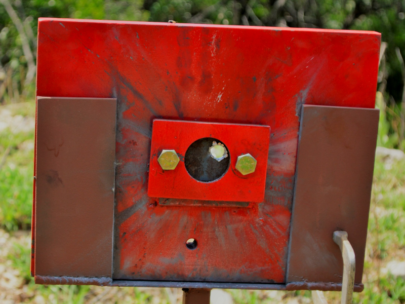 Red painted device where the 50 mm x 50 mm carbon steel plate was positioned (in the circle).  The image presents the target, positioned 100 meters far from the gun, after the shot and the projectile perforated the plate, as will be presented in the other figures.
