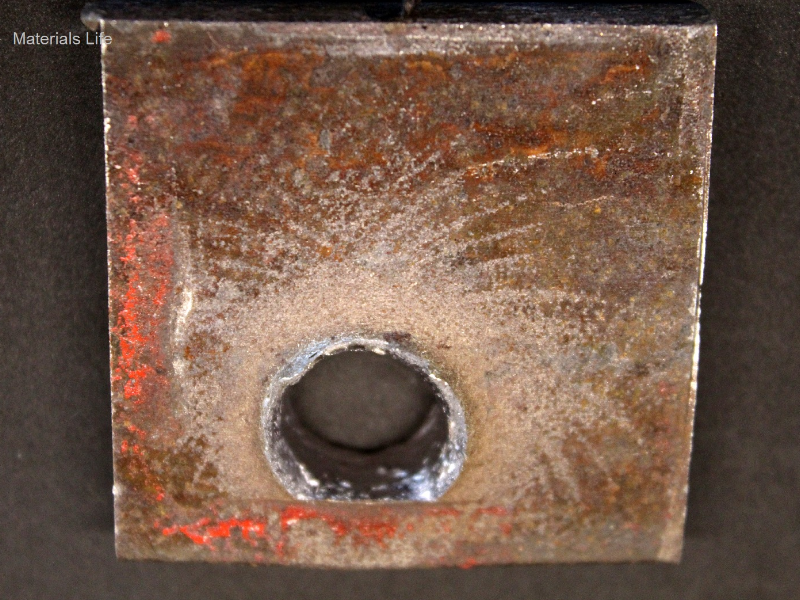 Other view of the 3/8" thickness plate, 50 mm x 50 mm, of the low carbon steel plate, in the opposite face of impact. The plate suffered a localized perforation and this image shows the position of the exit of the bullet. It is also evident that the shot was very close to the red painted device since a straight line is marked at the surface of the plate (bottom of the figure).