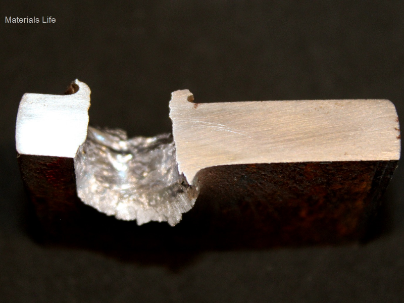 Aspect of the 3/8" thickness plate, 50 mm x 50 mm, of the low carbon steel, in a section transverse to the impact position that is to the top of the figure. The perforated hole expulsed the material to the top and bottom directions. The hole is larger in the exit of the bullet.