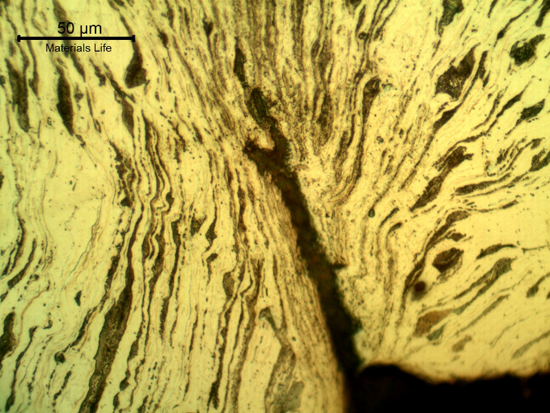 Micrographic detail of the damaged plate at the side of the bullet exit, bottom of the figure. The perforated hole is in the right side of the figure and it is evident a strong plastic deformation of the plate microstructure and microcracks.  In this region (exit of the bullet) there is no melted film anymore.