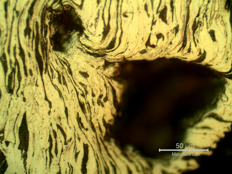 Micrographic detail of the damaged plate at the side of the bullet exit, bottom of the figure. The perforated hole is in the right side of the figure and it is evident a strong plastic deformation of the plate microstructure and microcracks.  In this region (exit of the bullet) there is no melted film anymore.