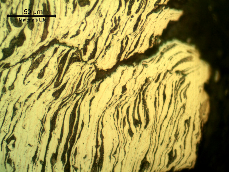 Micrographic detail of the damaged plate at the side of the bullet exit, bottom of the figure. The perforated hole is in the right side of the figure and it is evident a strong plastic deformation of the plate microstructure and microcracks.  In this region (exit of the bullet) there is no melted film anymore.