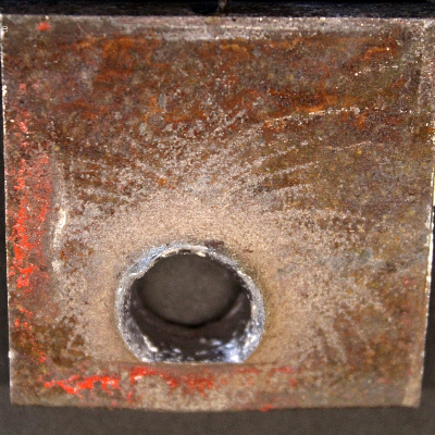 Other view of the 3/8" thickness plate, 50 mm x 50 mm, of the low carbon steel plate, in the opposite face of impact. The plate suffered a localized perforation and this image shows the position of the exit of the bullet.  It is also evident that the shot was very close to the red painted device since a straight line is marked at the surface of the plate  (bottom of the figure).
