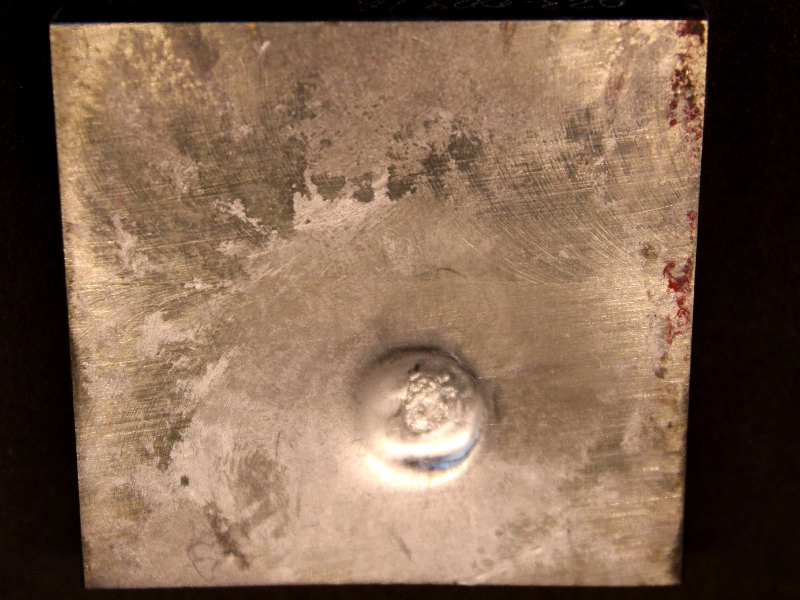 Other view of the 3/8" thickness plate, 50 mm x 50 mm, of titanium alloy, in the face of projectile impact. The plate suffered a localized plastic deformation, but no perforation.