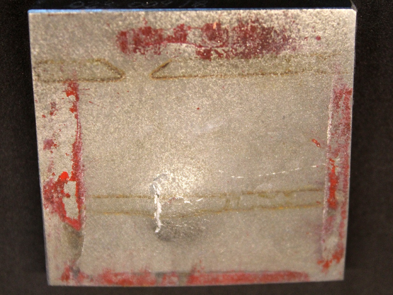 Other view of the 3/8" thickness plate, 50 mm x 50 mm, of the titanium alloy, in the opposite face of impact. The plate did not suffered perforation but it is fully cracked at the bullet impact. The  red painting of the device marked the back surface of the plate where it was in contact.