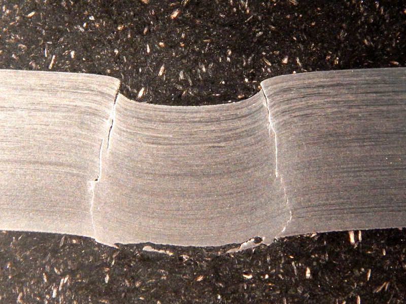 Macrography of the 3/8" thickness titanium alloy plate, in a section. The side of bullet impact is to the top of the figure. The impact sheared the plate wall in a very localized position. HF+HNO3+H2O chemical etch.