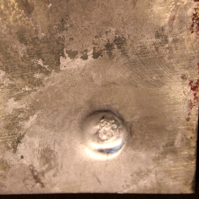 Other view of the 3/8" thickness plate, 50 mm x 50 mm, of titanium alloy, in the face of projectile impact. The plate suffered a localized plastic deformation, but no perforation.