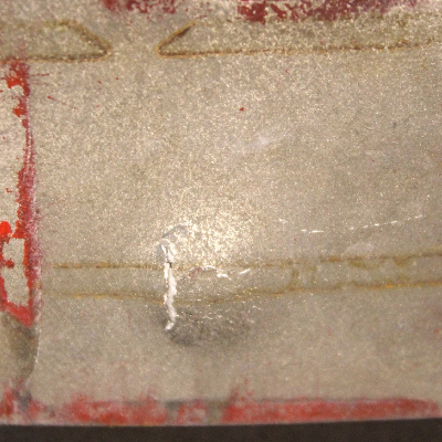 Other view of the 3/8" thickness plate, 50 mm x 50 mm, of the titanium alloy, in the opposite face of impact. The plate did not suffered perforation but it is fully cracked at the bullet impact. The red painting of the device marked the back surface of the plate where it was in contact.