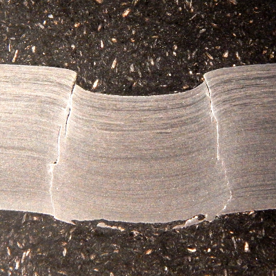 Macrography of the 3/8" thickness titanium alloy plate, in a section. The side of bullet impact is to the top of the figure. The impact sheared the plate wall in a very localized position. HF+HNO3+H2O chemical etch.