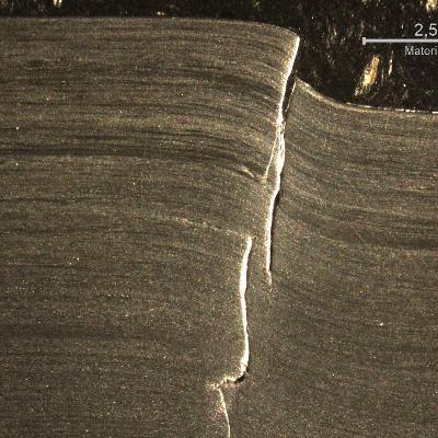 Macrography of the 3/8" thickness titanium alloy plate, in a section. The side of bullet impact is to the top of the figure. The impact sheared the plate wall in a very localized position.  HF+HNO3+H2O chemical etch.