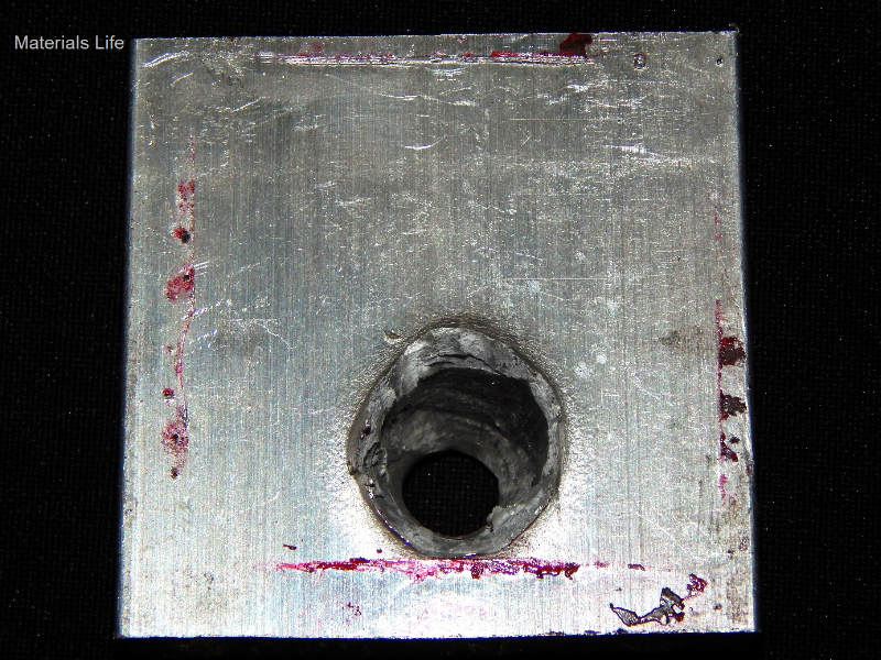Other view of the 3/8" thickness, 50 mm x 50 mm, 6061 T6 aluminum plate, in the opposite face of impact. The plate suffered a localized perforation and this image shows the position of the exit of the bullet.