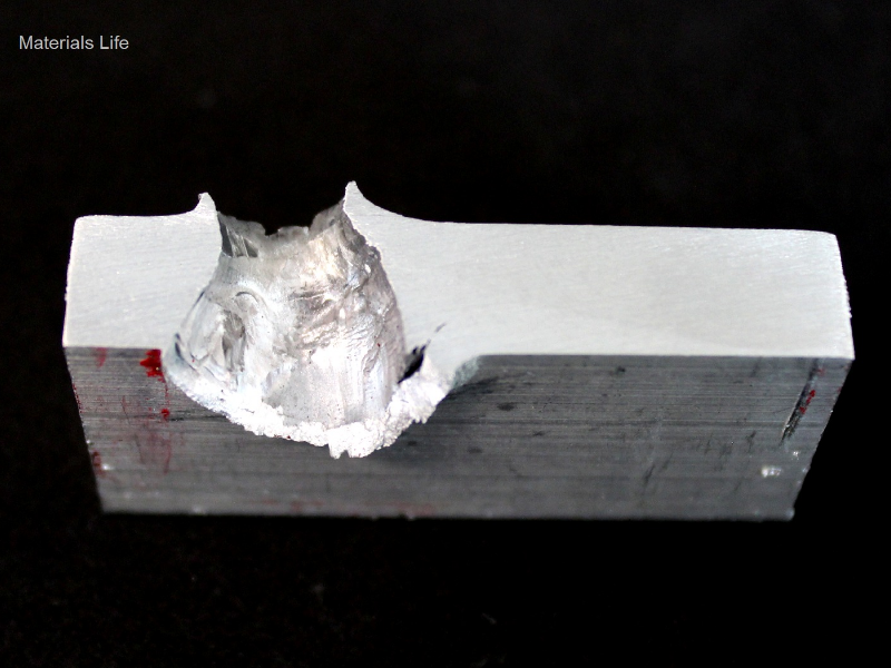 Aspect of the 3/8" thickness, 50 mm x 50 mm, 6061 T6 aluminum plate, in a section transverse to the impact position that is to the top of the figure. The perforated hole expulsed the material to the top and bottom directions. The hole is larger in the exit of the bullet.
