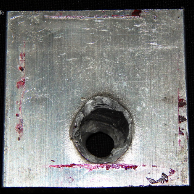 Other view of the 3/8" thickness, 50 mm x 50 mm, 6061 T6 aluminum plate, in the opposite face of impact. The plate suffered a localized perforation and this image shows the position of the exit of the bullet.