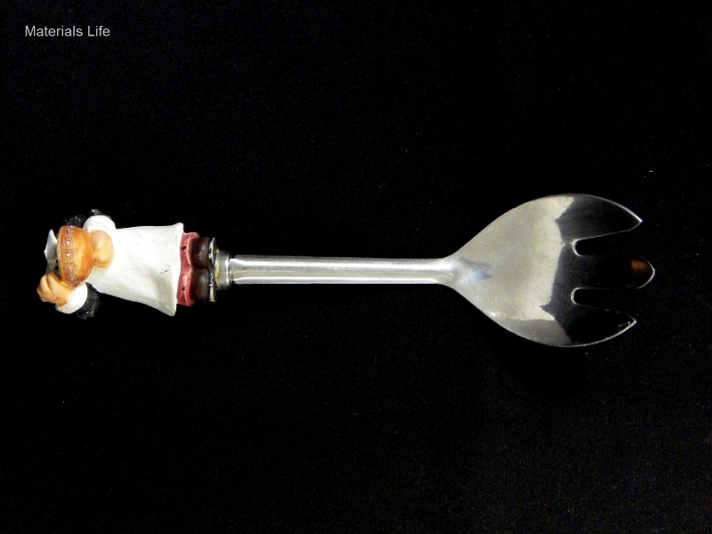 Decorative table service fork, with a non metallic handle that is broken. The metallic part is about 165 mm long and presents two shapes: the scoop and the handle. The scoop is 53mm wide and 73mm long and the handle is about 9 mm wide and 75 mm long.