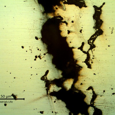 Cross-sectional micrograph showing a wide, irregular crack with branching dark deposits through the polished matrix, alongside scattered small voids.